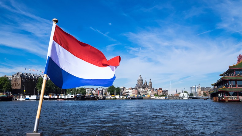 Flag of the Netherlands flies from the back of a boat, with canal water and buildings behind
