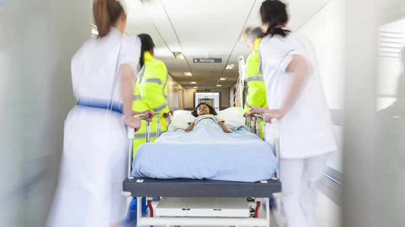 A motion blurred photograph of a young Asian Indian girl child patient on stretcher or gurney being pushed at speed through a hospital corridor by doctors & nurses to an emergency room - Image 