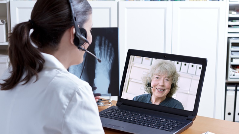 Female doctor in her surgery office with headset in front of her laptop, an x-ray of a foot in hand, talking with a senior patient, telemedicine concept