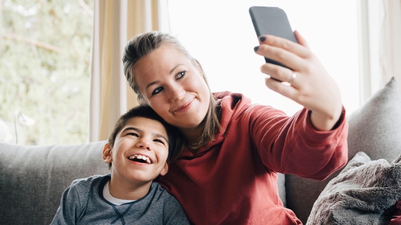 Smiling mother taking selfie with autistic son while sitting on sofa at home