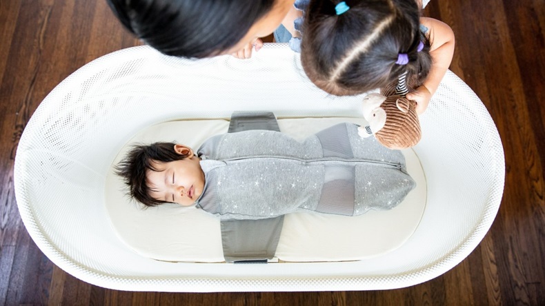 A family gathers around a dark-haired infant asleep in a SNOO bassinet. 