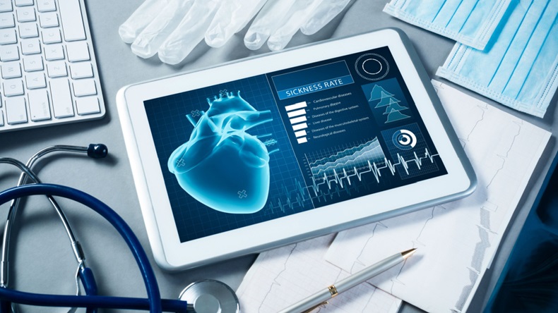 An ipad on a desk with a digital image of a heart. Also pictured slightly out of frame are a keyboard, medical gloves, a pen and paper, surgical masks, and a stethescope