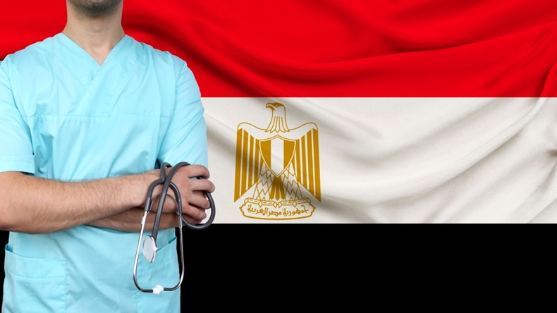 Medical professional in blue scrubs standing in front of the Egyptian flag.